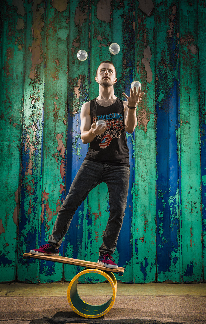 Colour photographic portrait of male juggler on wobble board juggling clear galss 'balls'.